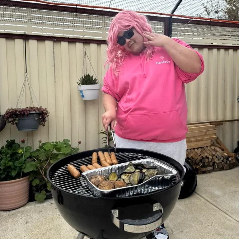 Me in a long pink wig, sunglasses and a pink HeadsUp hoodie, posing with tongs over a kettle barbecue of sausages and grilled eggplant
