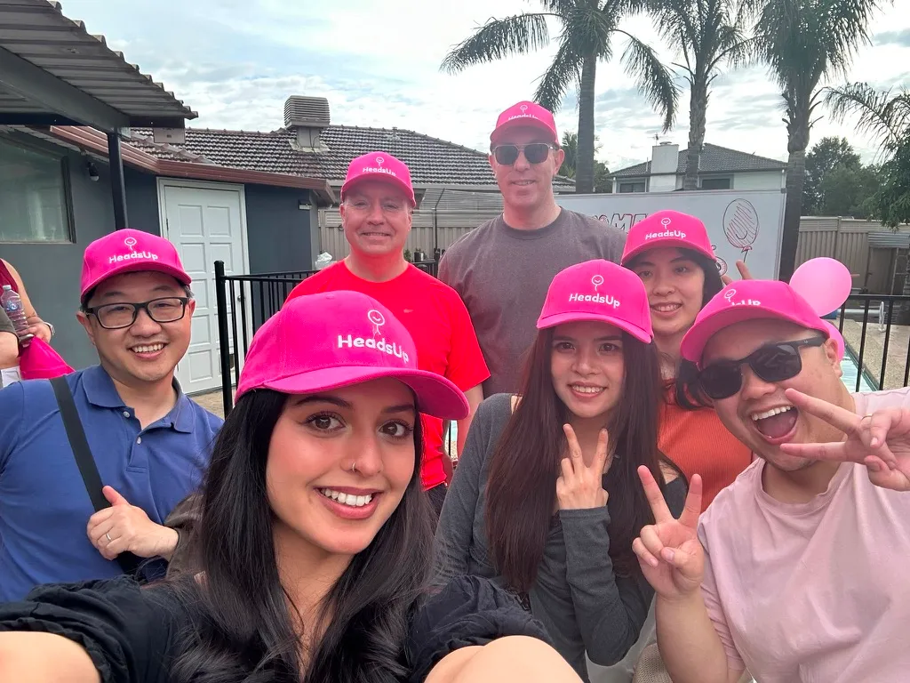 A selfie of seven friends grinning and throwing peace signs, all wearing pink HeadsUp caps